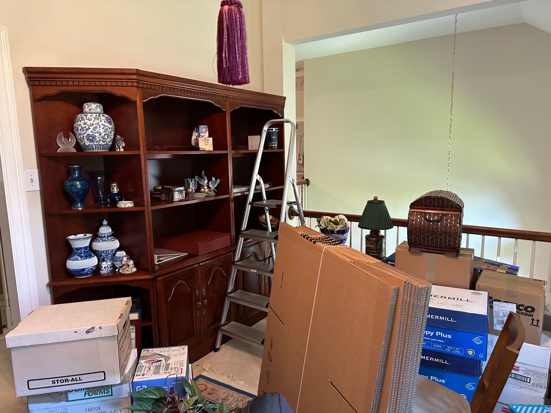 A wooden bookshelf filled with ceramics next to a ladder, boxes, and a table in a room during a move or renovation.