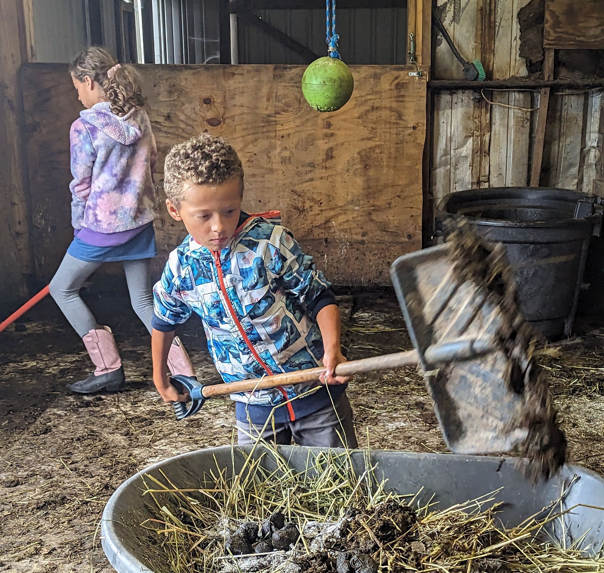 a girl and a boy next to a wheelbarrow and mucking a stall