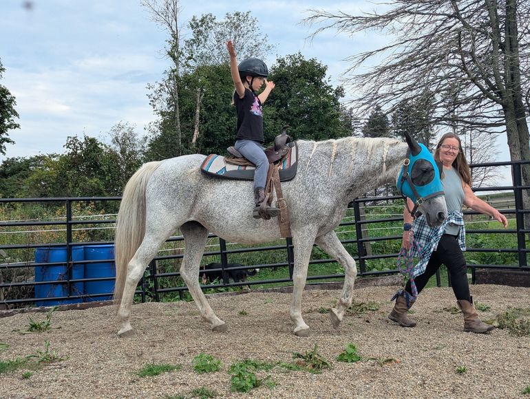 Child riding a white horse while another person leads it in a paddock, with barrels and trees nearby