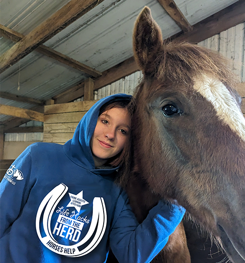 Young lady, wearing a blue hoodie with LIFE HACKS FROM THE HERD logo, poses with a horse