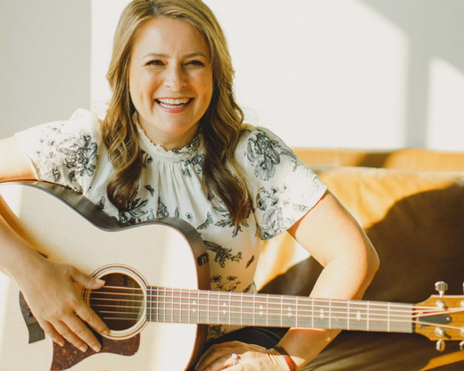Melanie Waldman: AUG 21st @7-9pm Person smiling and playing acoustic guitar on a couch in a sunlit room