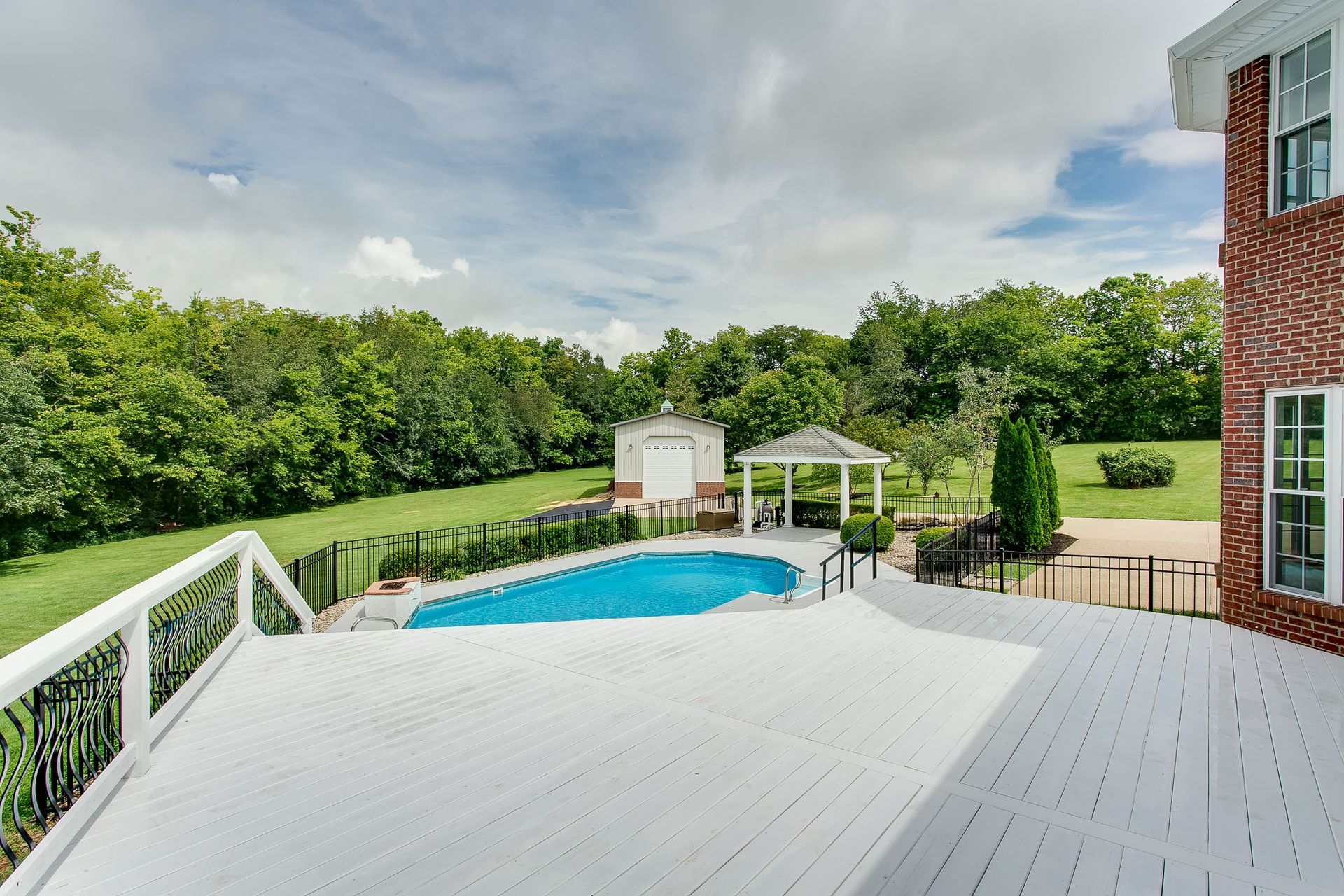 White deck overlooking a backyard pool, gazebo, and green trees under a cloudy sky