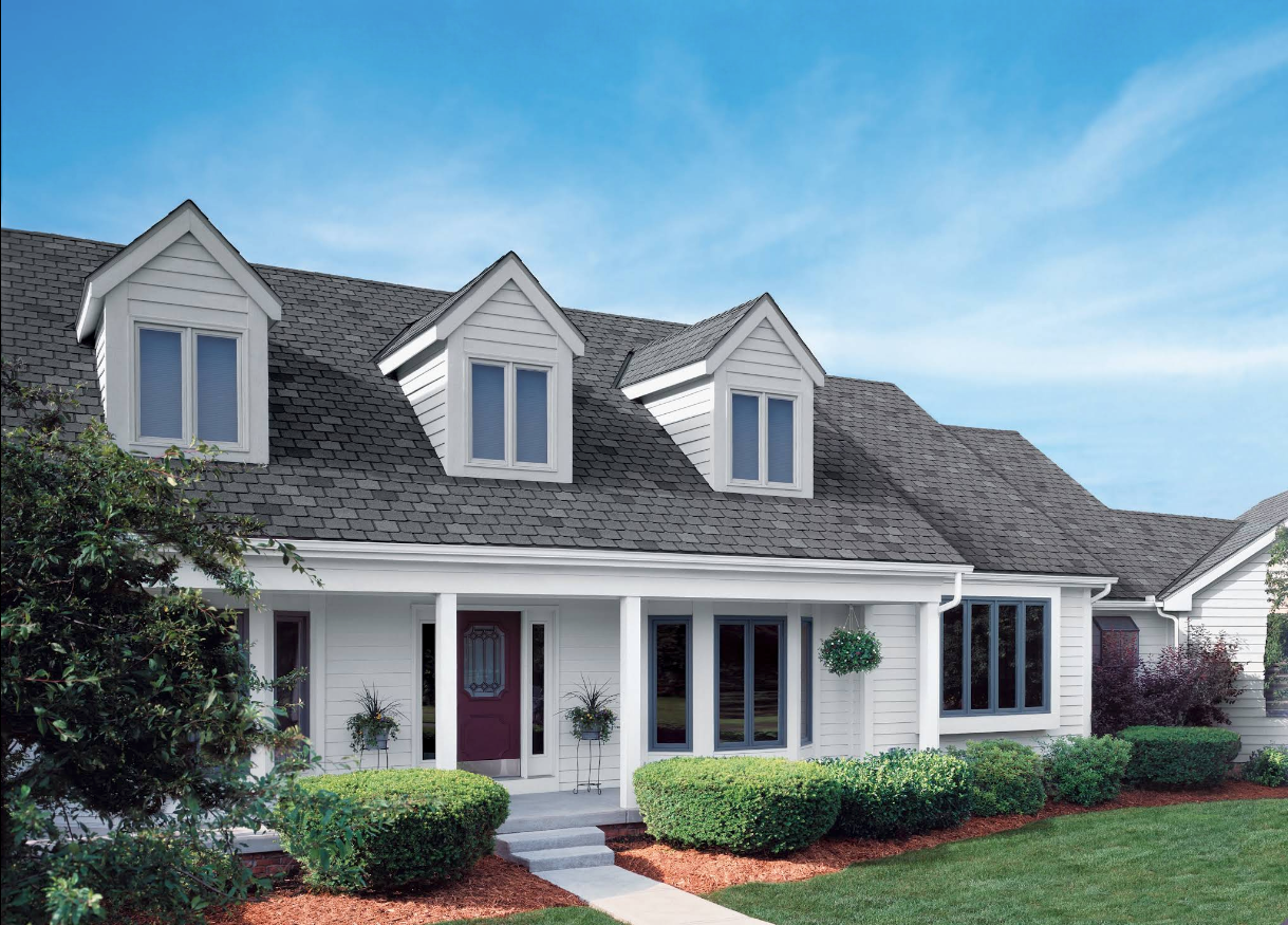 White suburban house with gray shingle roof, dormer windows, white porch, and red front door under a blue sky