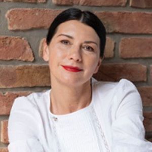 A woman wearing a white shirt and red lipstick is standing in front of a brick wall.