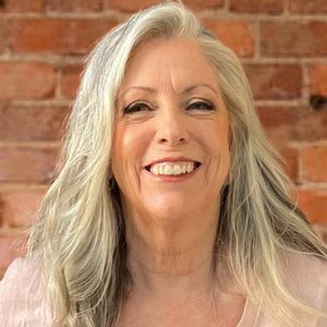 A woman with gray hair is smiling in front of a brick wall.