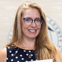 A woman wearing glasses and a polka dot dress is holding a piece of paper and smiling.