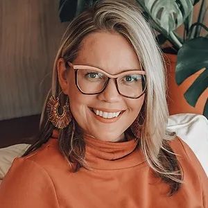 A woman wearing glasses and earrings is smiling while sitting on a bed.