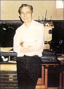 Man in white shirt and dark pants smiles, arms crossed, standing near machinery in a workshop.