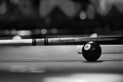 Close-up of a pool cue and 8-ball on a billiards table in black and white.