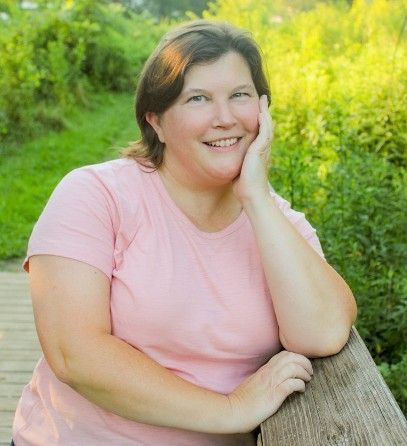 Woman in pink shirt smiles outdoors, resting chin on hand and arm on wooden railing. Green foliage in background.