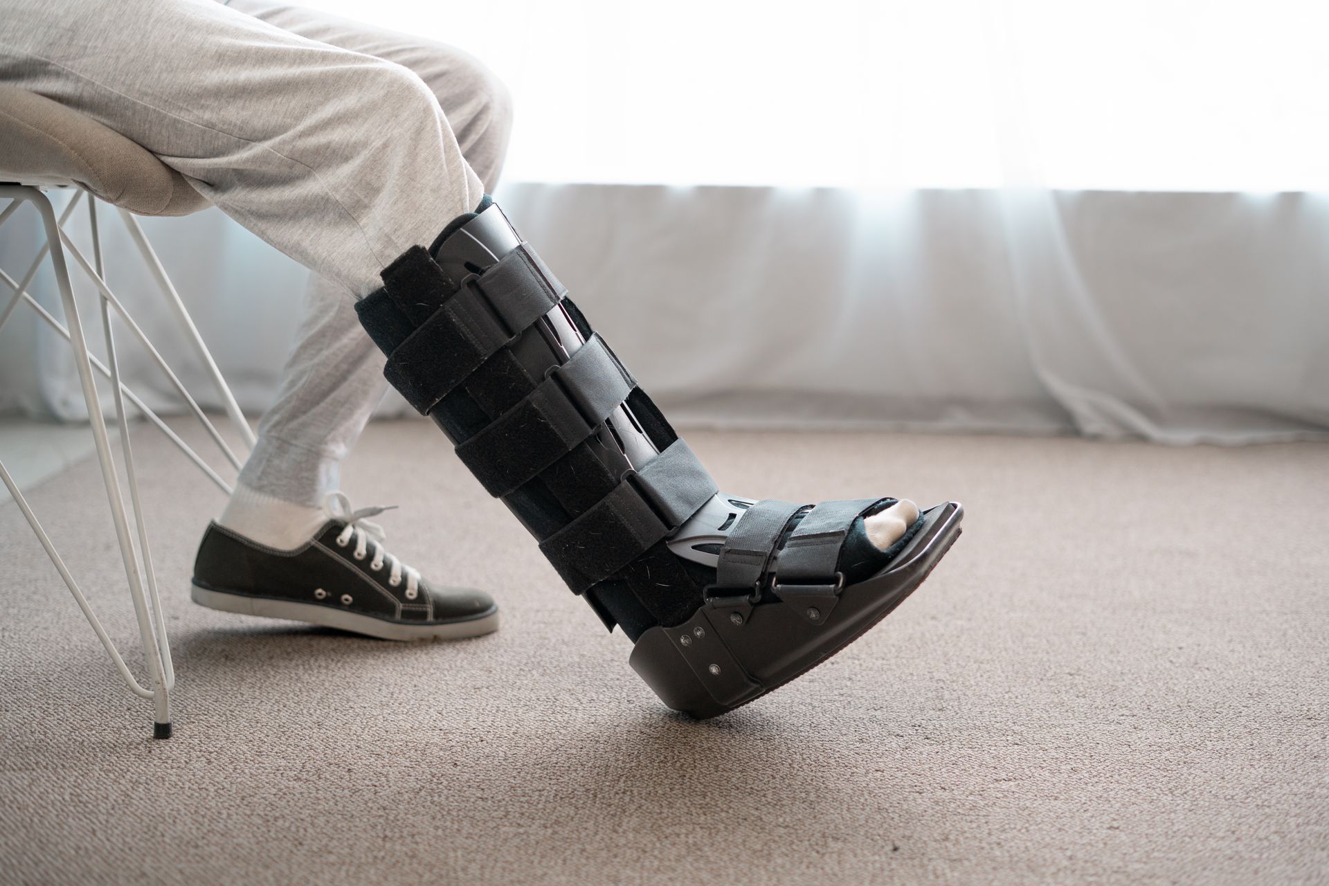 Person's leg in a black walking boot, other foot in a sneaker, sitting near a window.