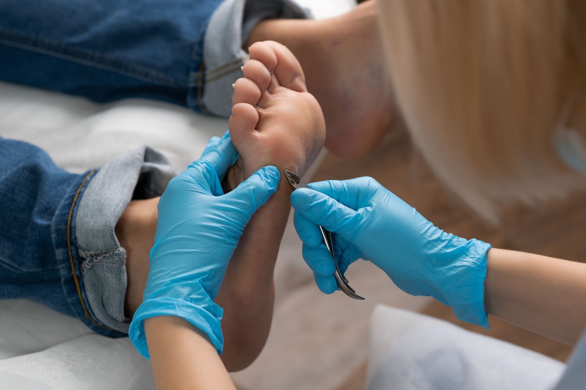 Person's foot being examined, close up. Person in blue gloves using a tool to examine the sole. Jeans visible.