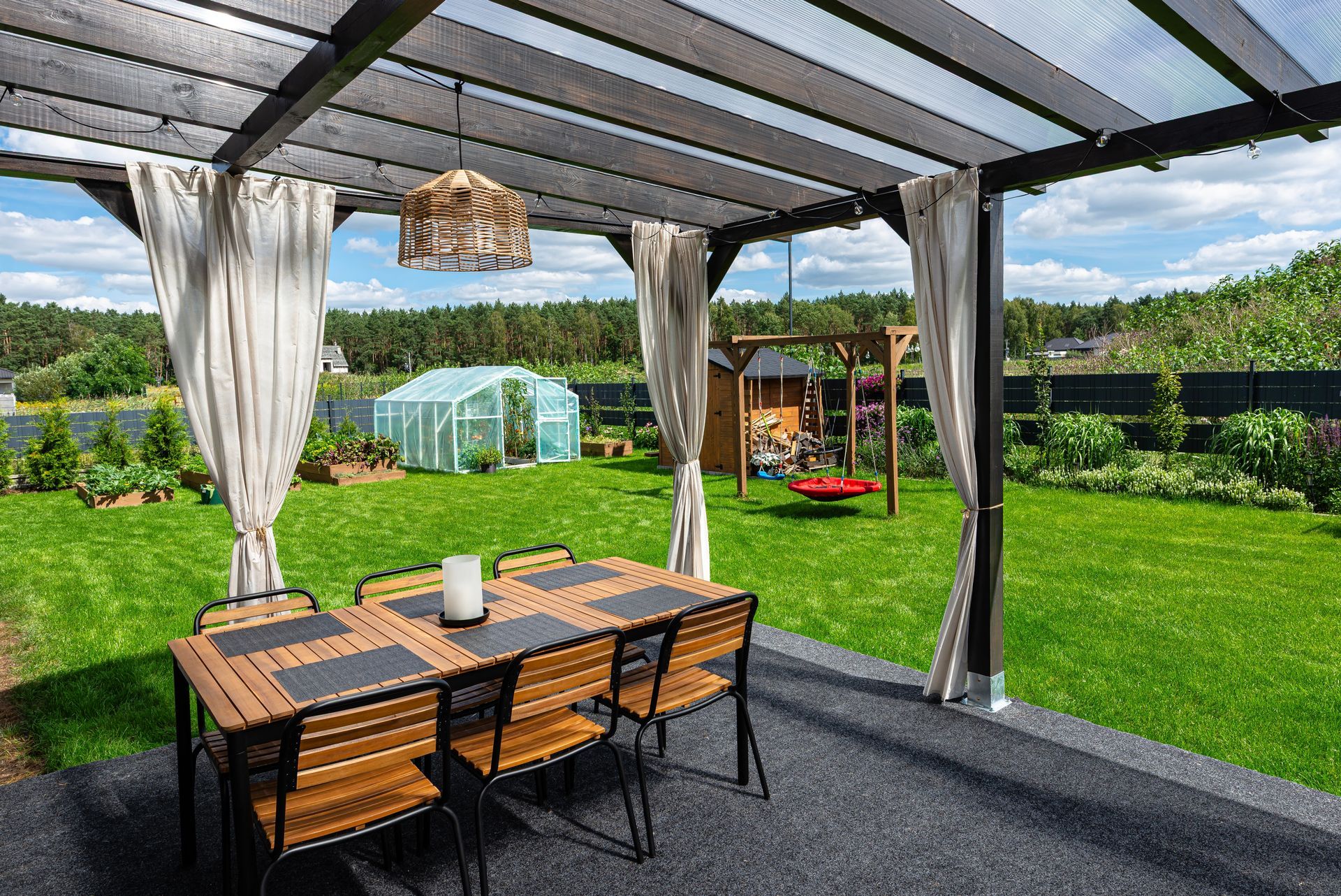 Covered patio with wooden dining table, curtains, and a sunny backyard garden with greenhouse and lawn.