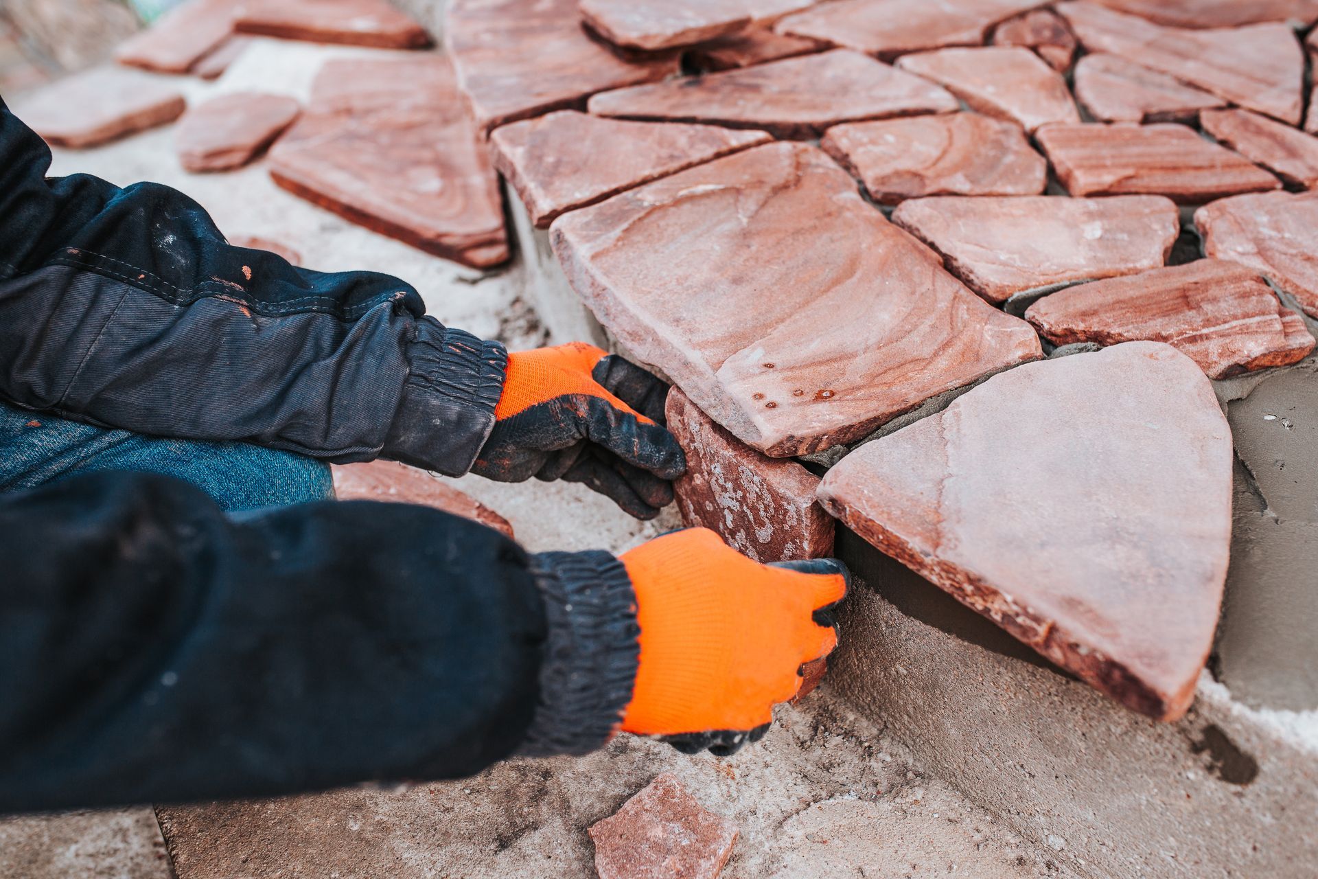 Orange-gloved hands laying stone pavers on a sandy surface