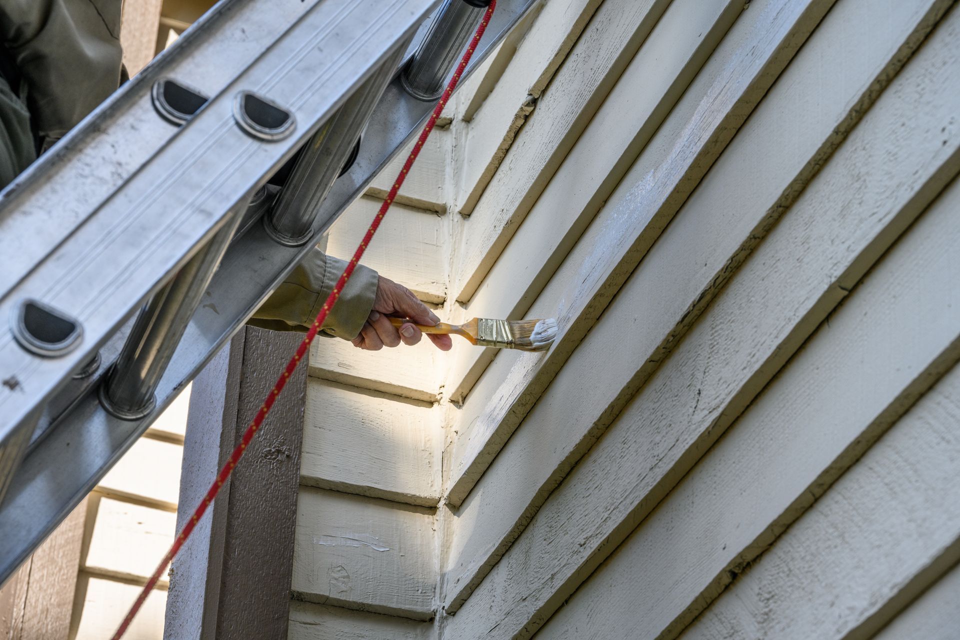 Worker on ladder using power drill on white house siding near roofline