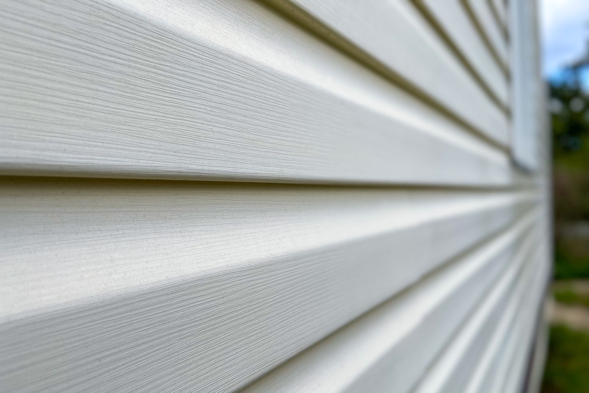 Close-up of white vinyl siding on a house, viewed at an angle with blue sky and greenery in the background