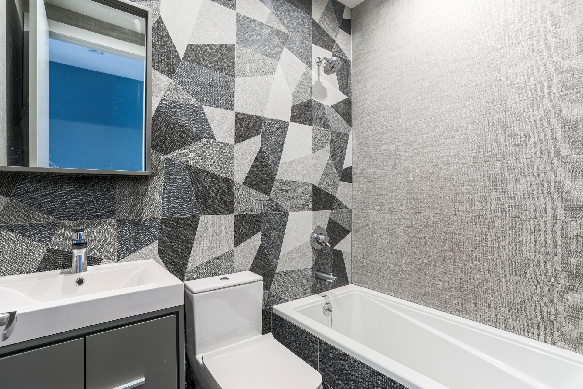 Small bathroom with geometric tile accent wall, white sink and toilet, and a bathtub with gray textured walls.