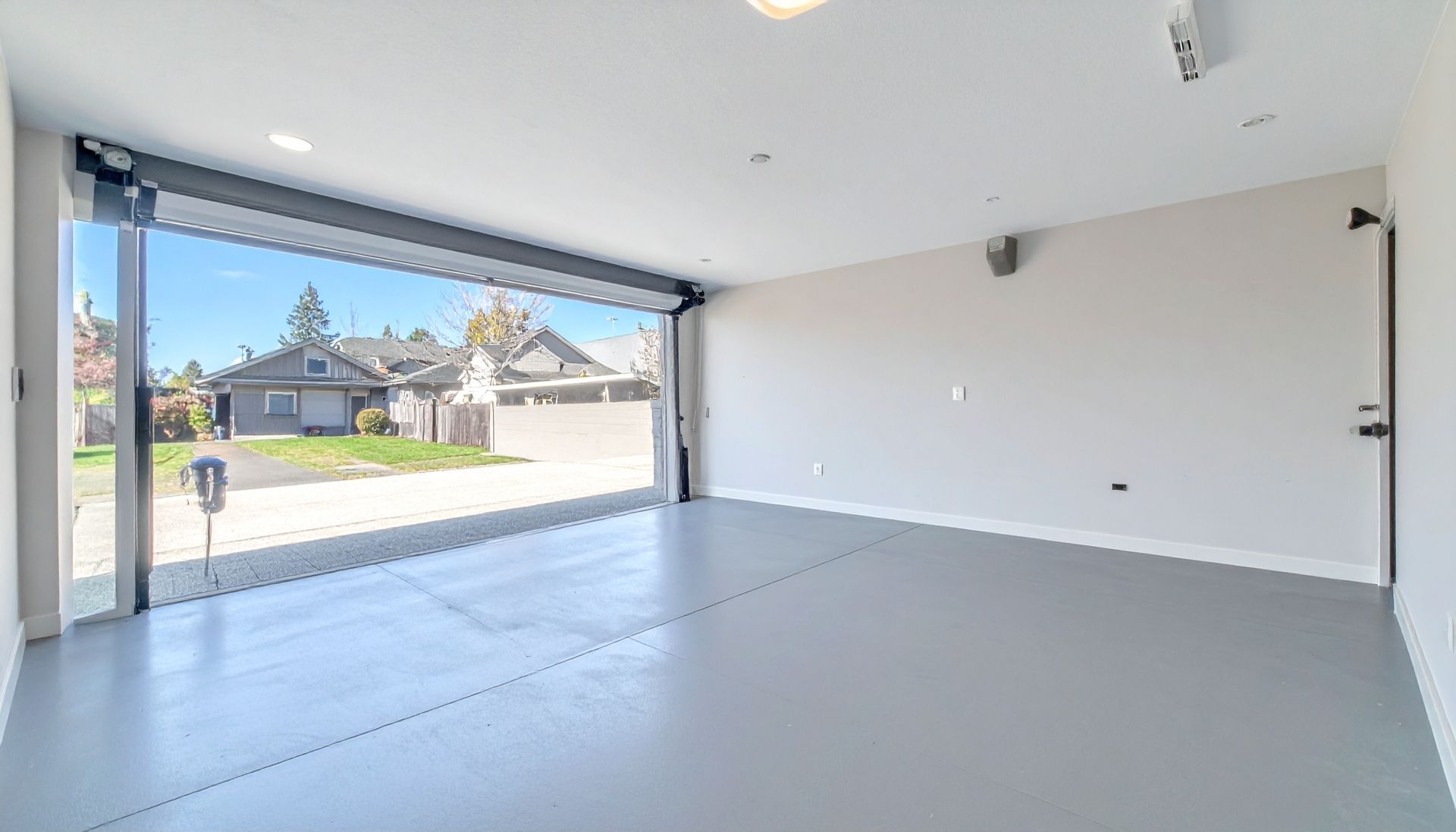 Bright empty garage with open door, white walls, gray floor, and view of a suburban street.