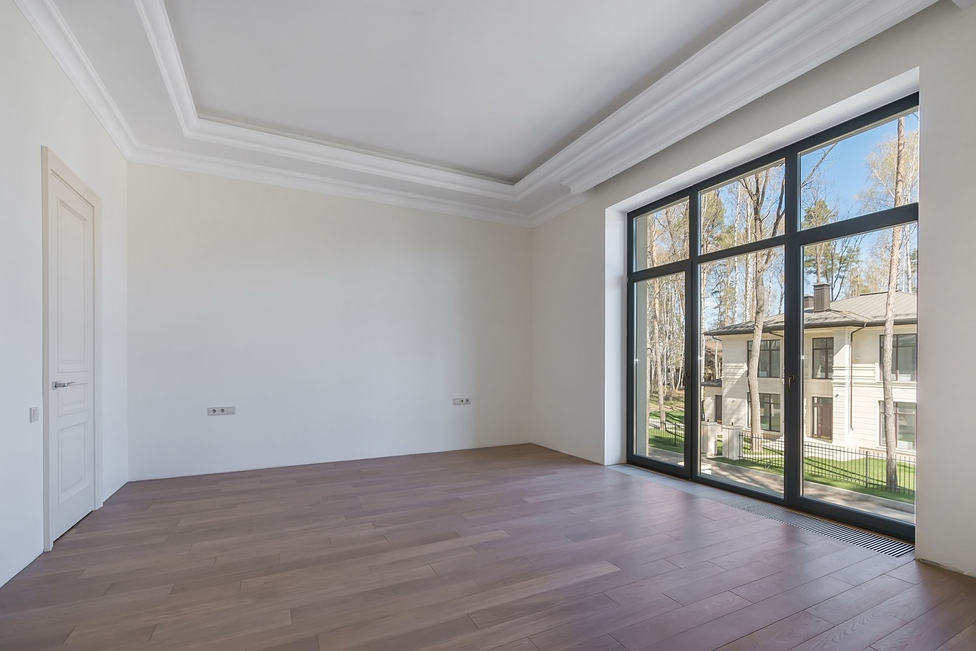 Empty modern room with light wood floors, white walls, and large black-framed windows overlooking greenery
