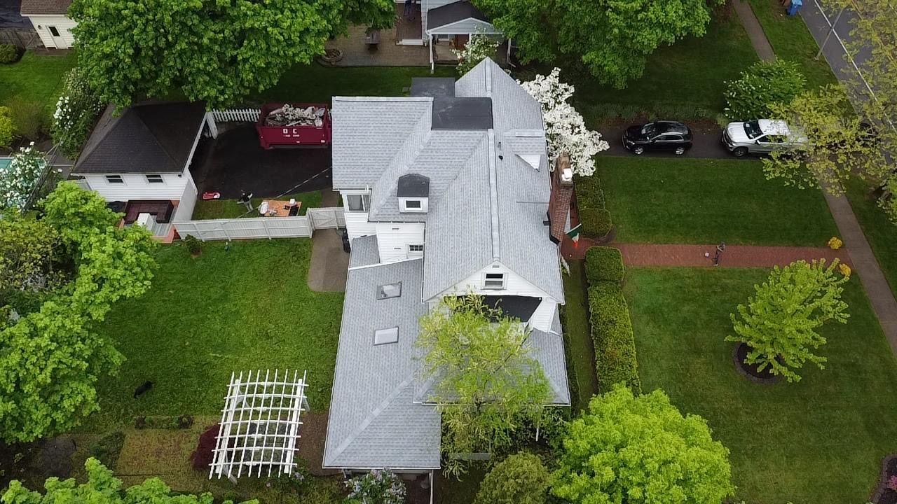 Aerial view of a house with a gray roof, surrounded by green grass and trees. Two cars parked on the street.