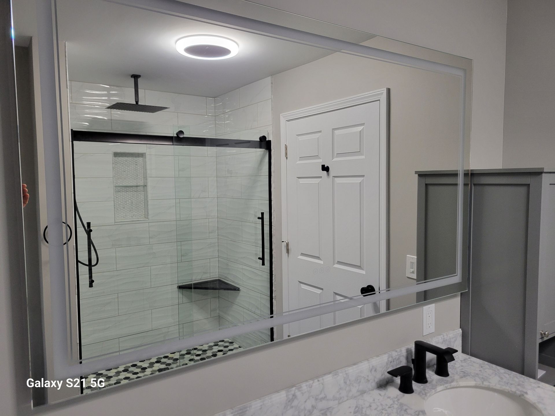 Bathroom mirror reflecting a shower, door, and sink. Black fixtures against white and gray decor.