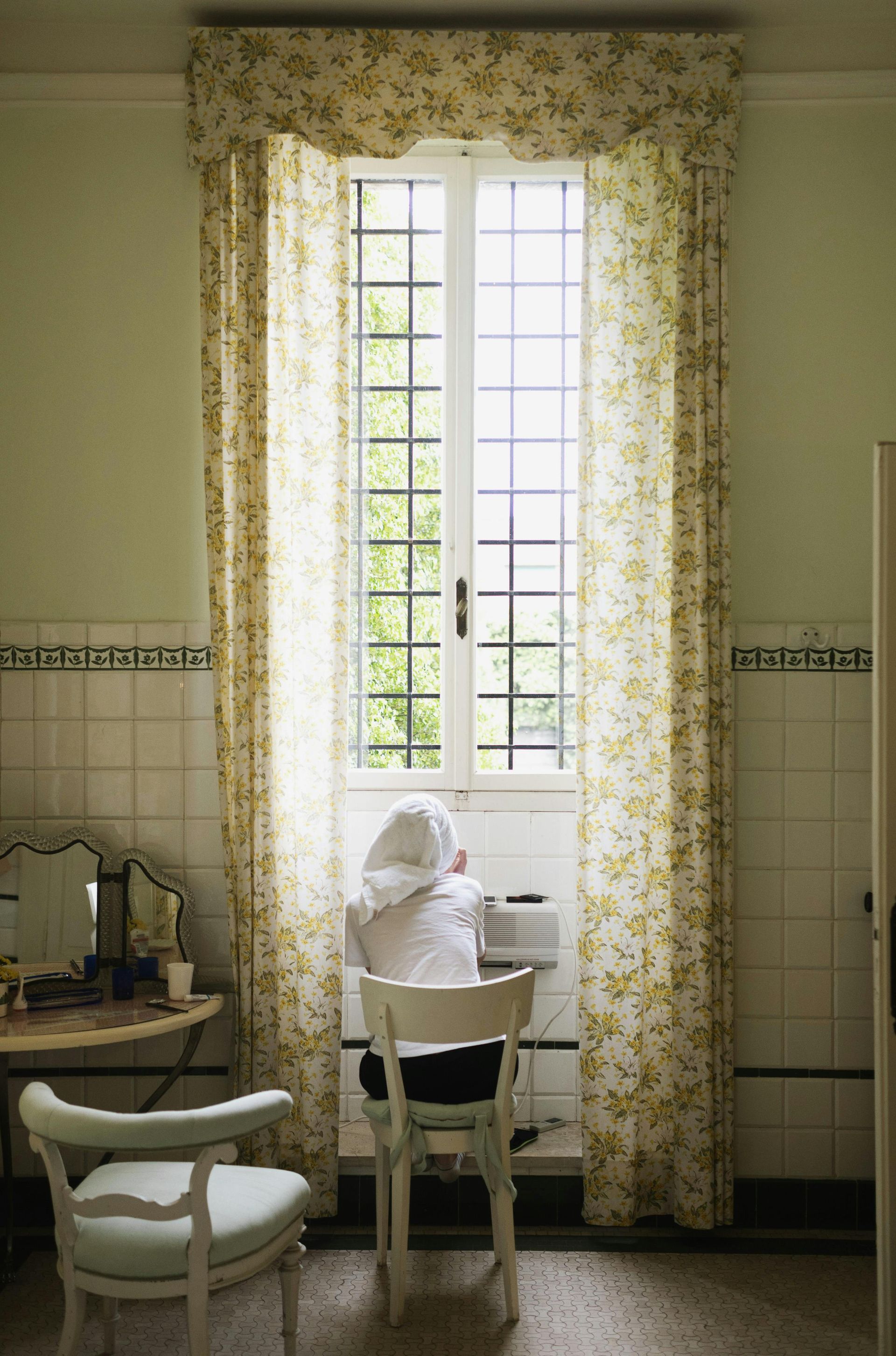 A person at a desk by a tall window with floral curtains. Green walls and a vanity mirror.