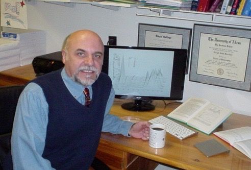 Man seated at a desk with a computer, papers, open book, and framed certificates in an office.