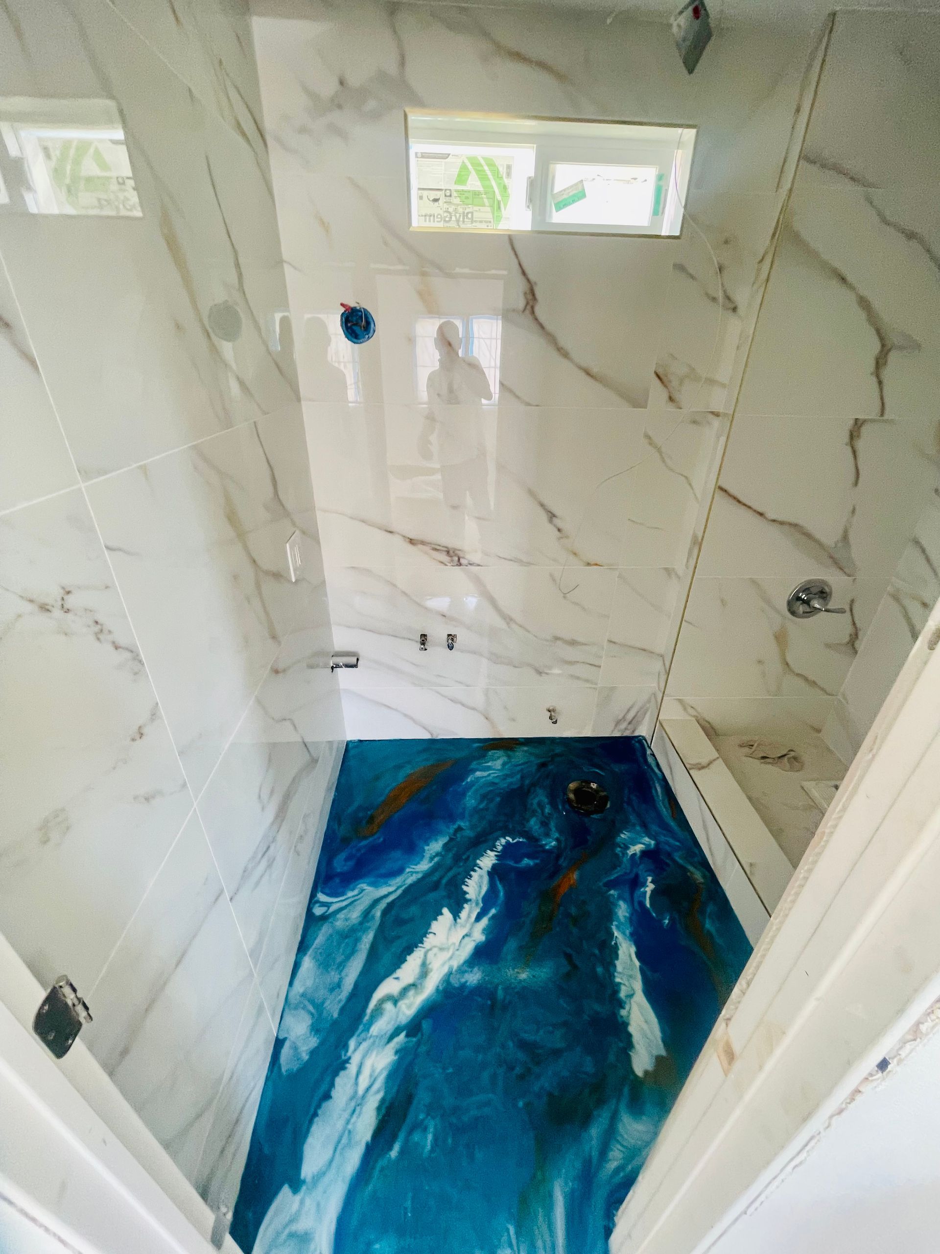 Blue and white epoxy bathroom floor with marble-tiled walls, two shower heads, and a small window.