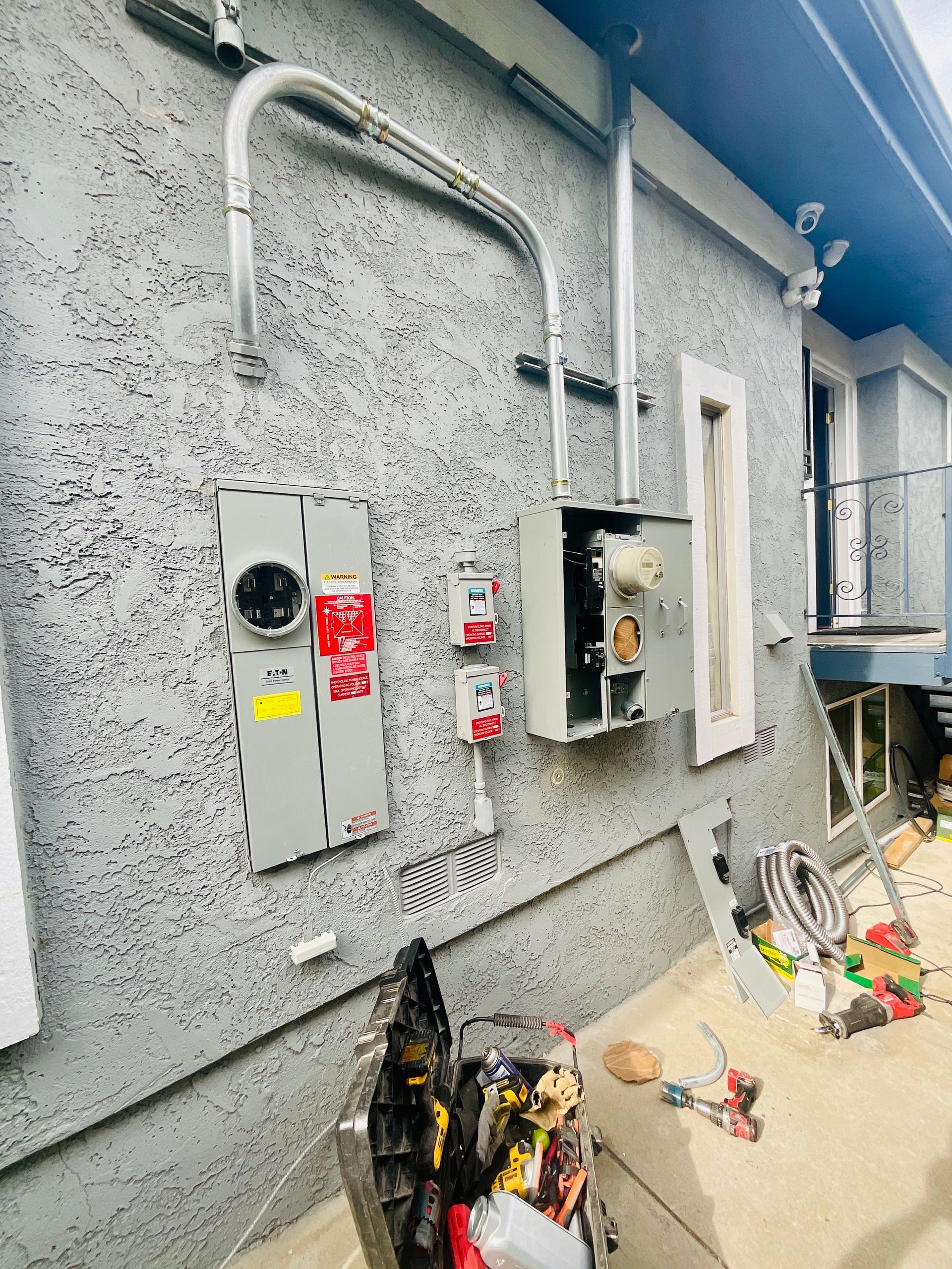 Exterior view of a house with electrical components: meter, panel, conduits, and junction boxes mounted on the wall.