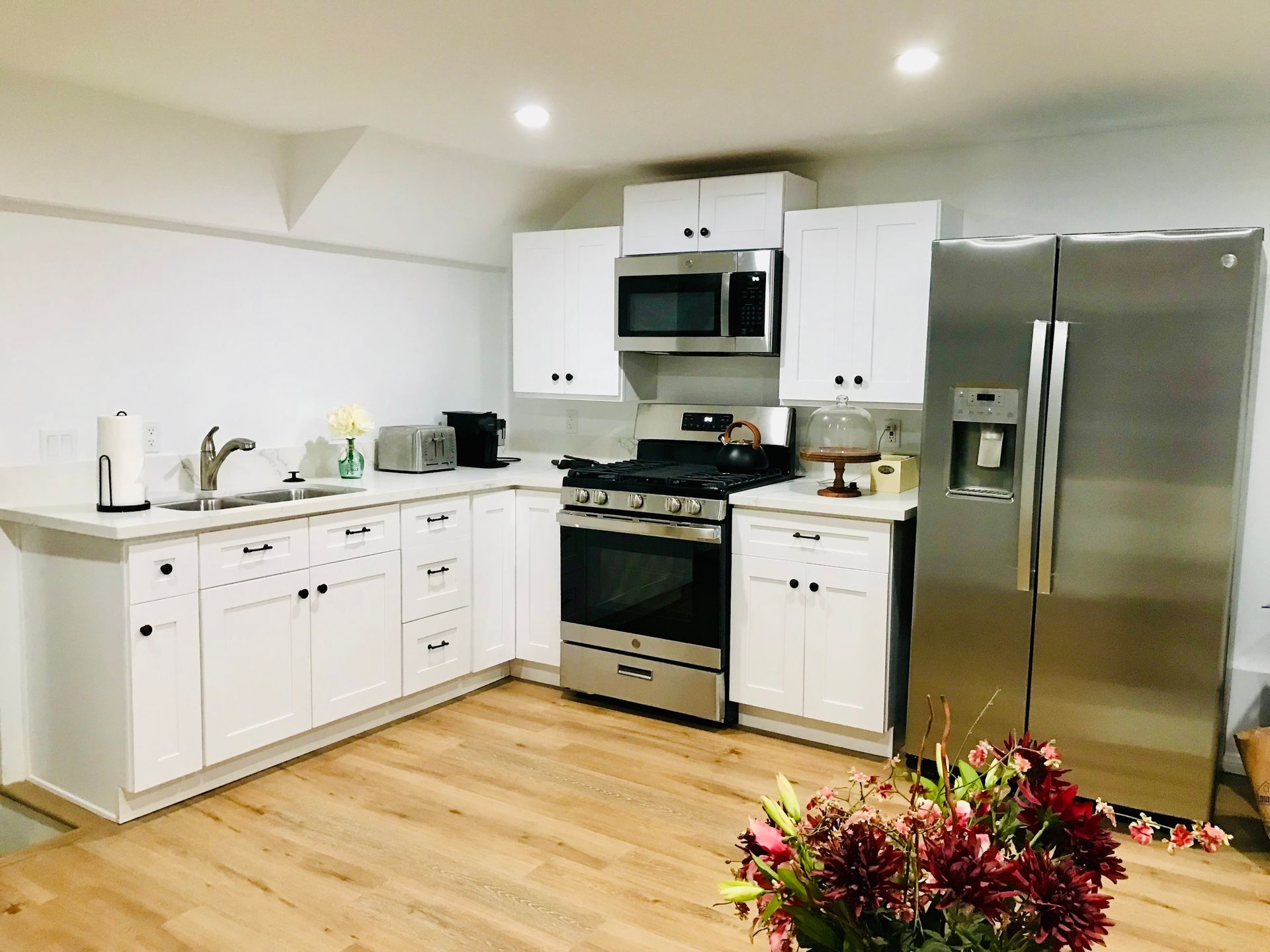 Modern white kitchen with stainless steel appliances, wooden floors, and flowers.