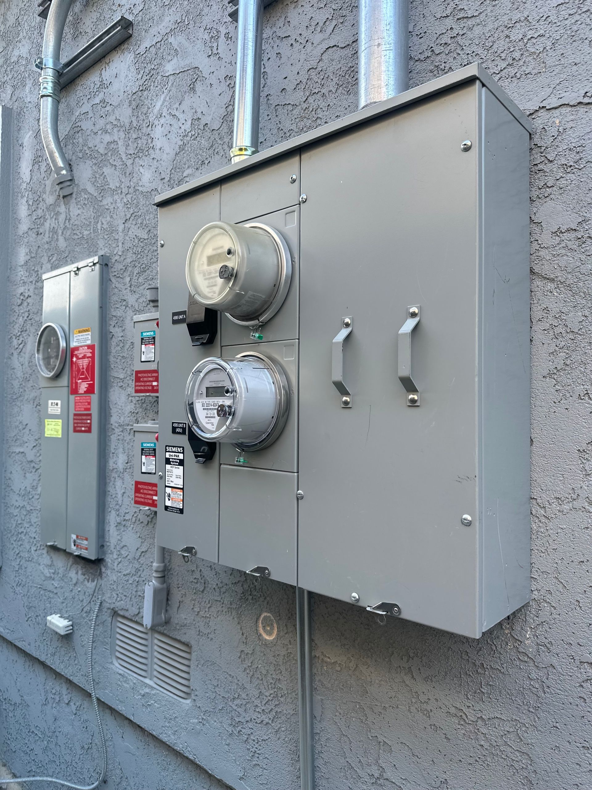 Gray electrical panel with two meters, mounted on a textured wall, with conduit.