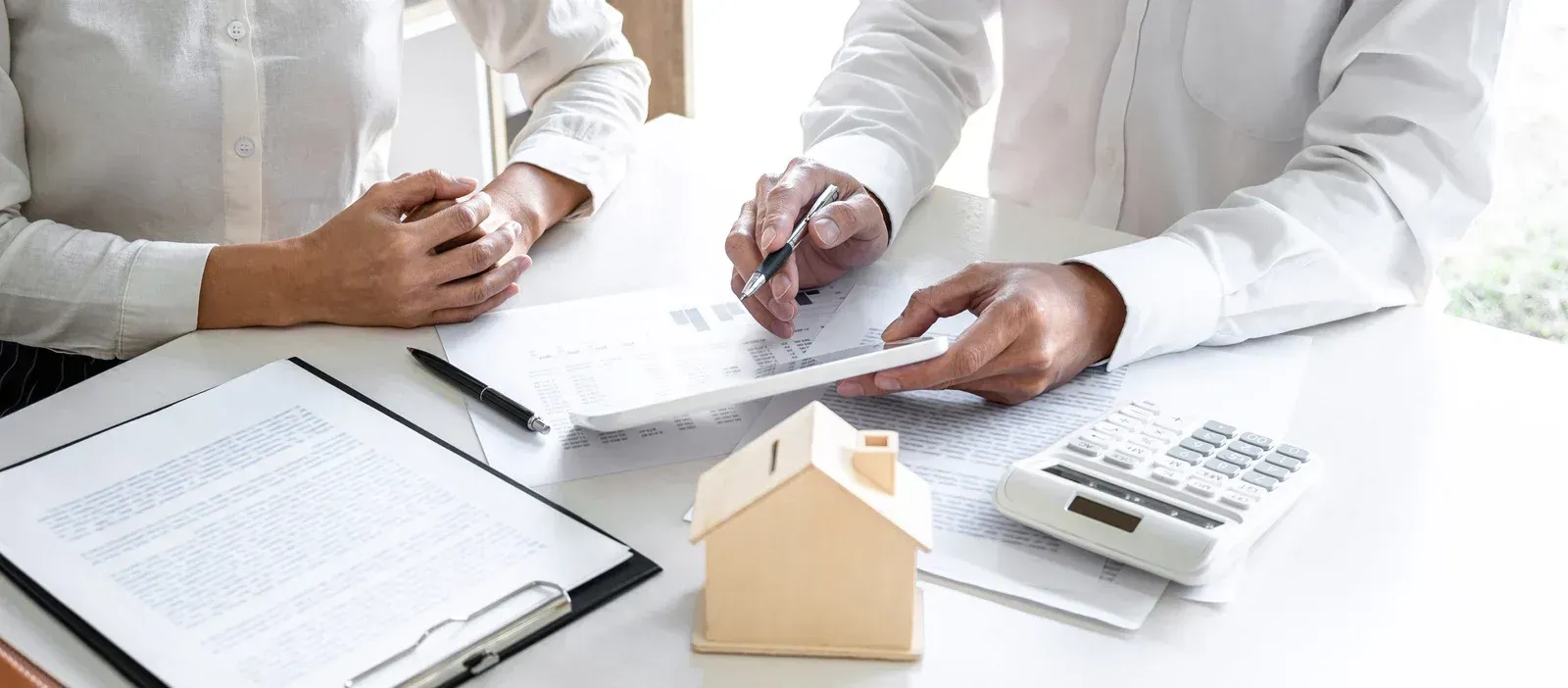 Two people review paperwork with a small house model and calculator on a desk.