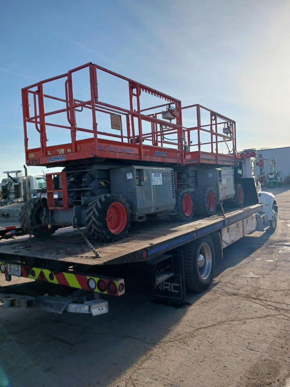 A large orange scissor lift loaded on a flatbed truck, outdoors. The lift is gray with orange wheels.