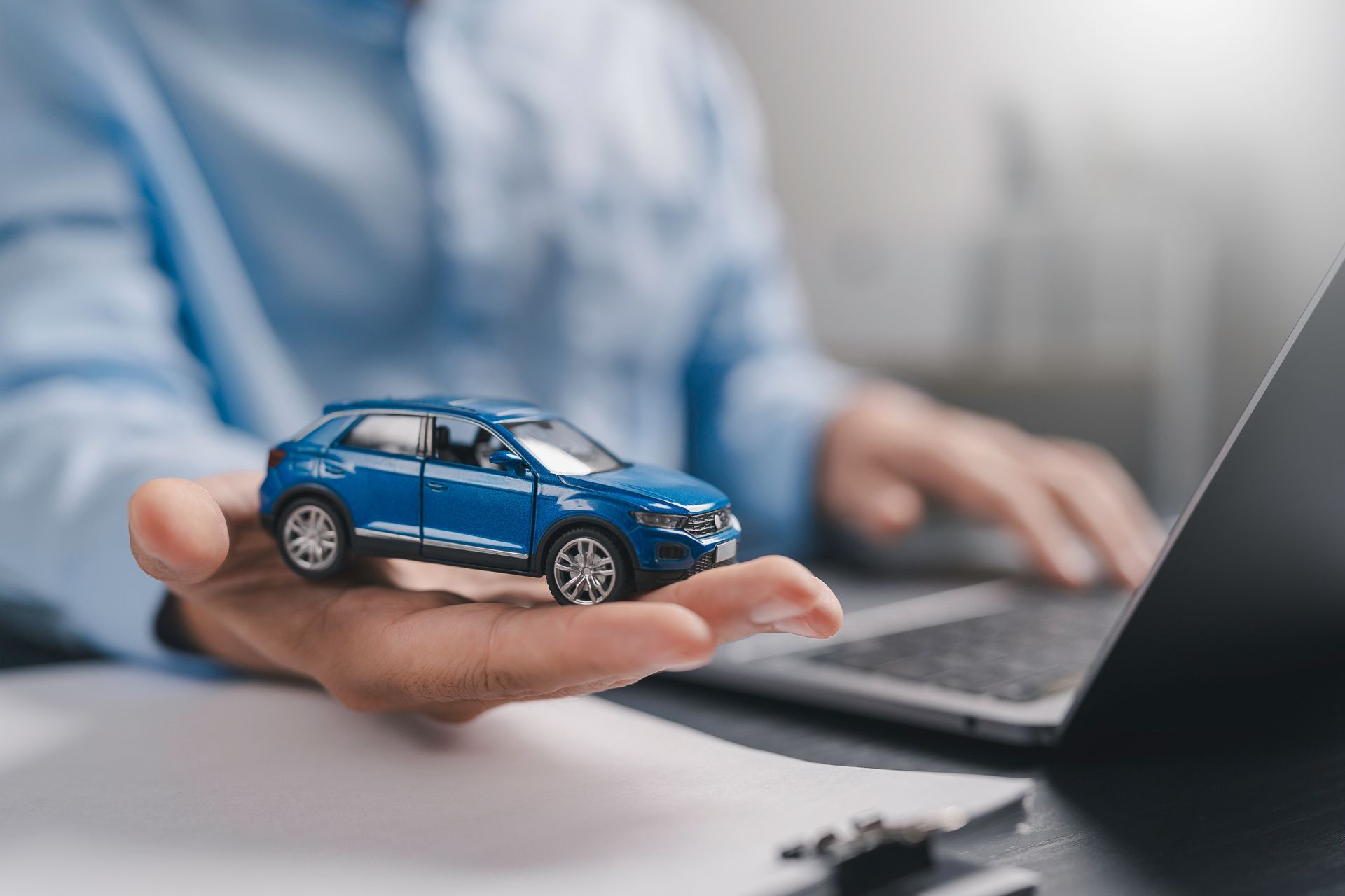Vehicle insurance agent holding a blue toy car while working on a laptop at a desk.