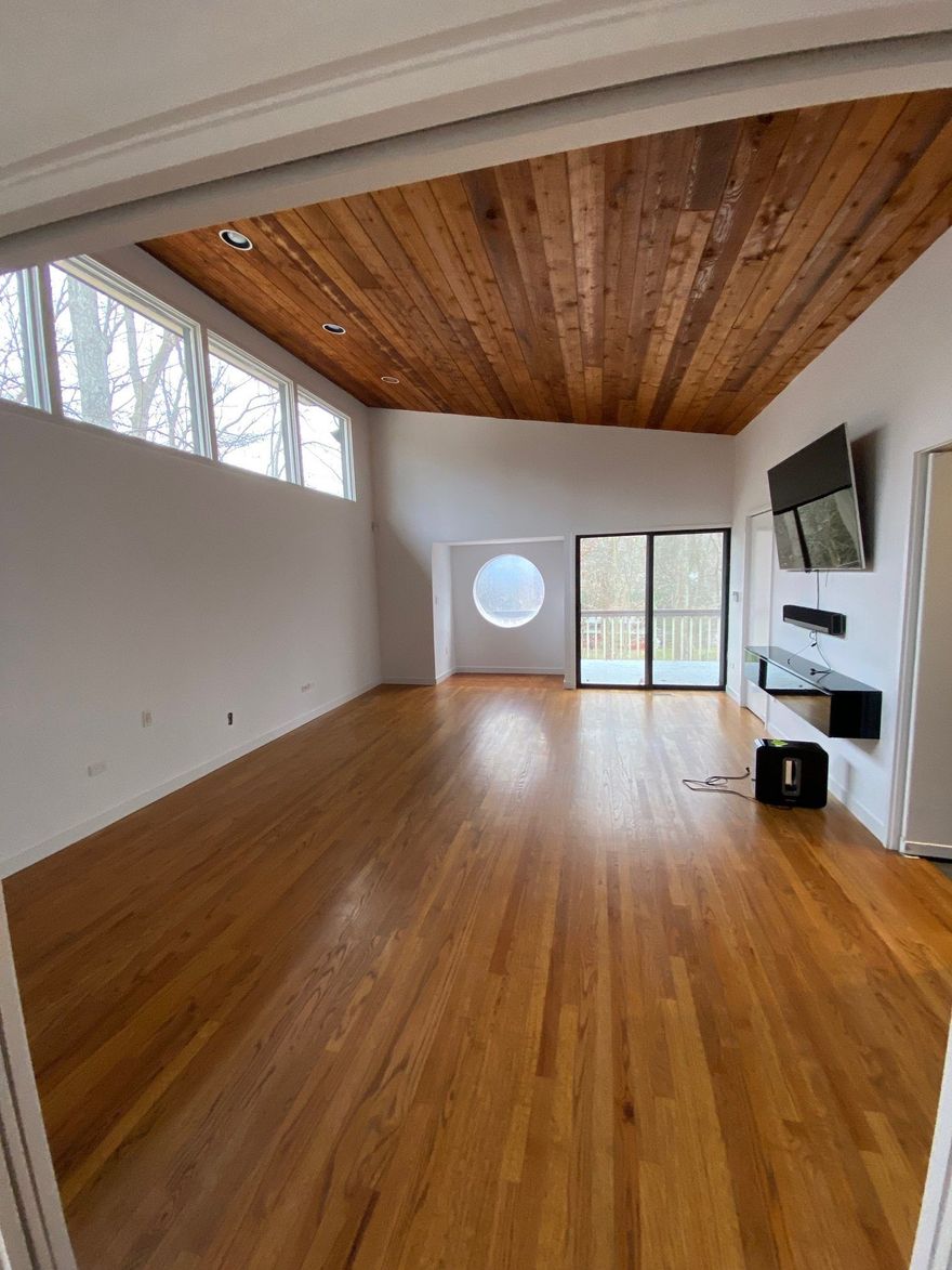 An empty, modern room with wooden floors, a matching paneled ceiling, large windows, a circular window, and a wall TV.