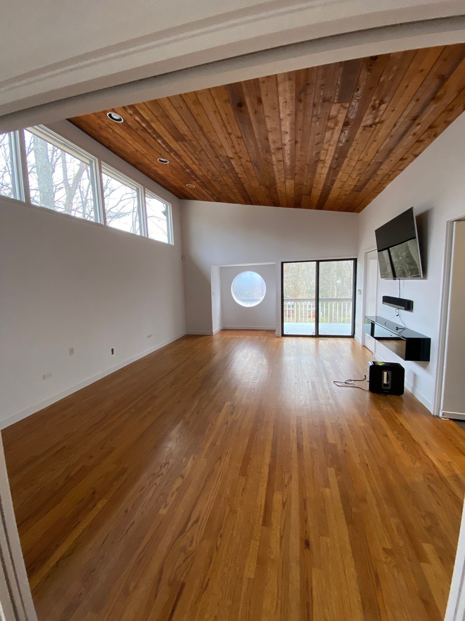 Empty modern living room with wood floors, sloped wood ceiling, large windows, fireplace, and wall-mounted TV