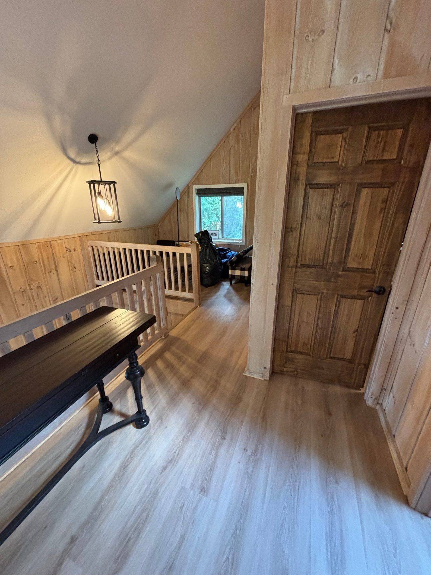 A hallway with light wood paneling and flooring, a wooden door, a black console table, and an attic window in the distance.