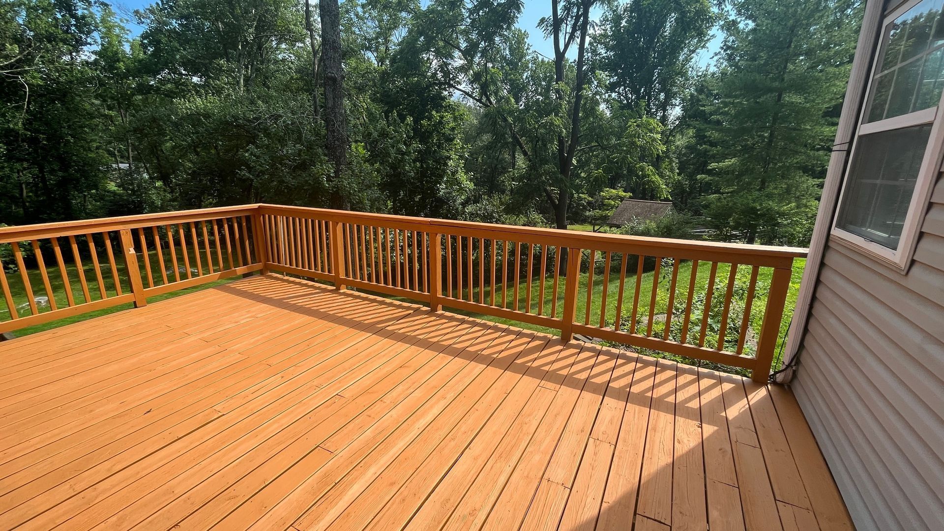 A wooden deck with a railing overlooks a lush green yard surrounded by trees under a blue sky.