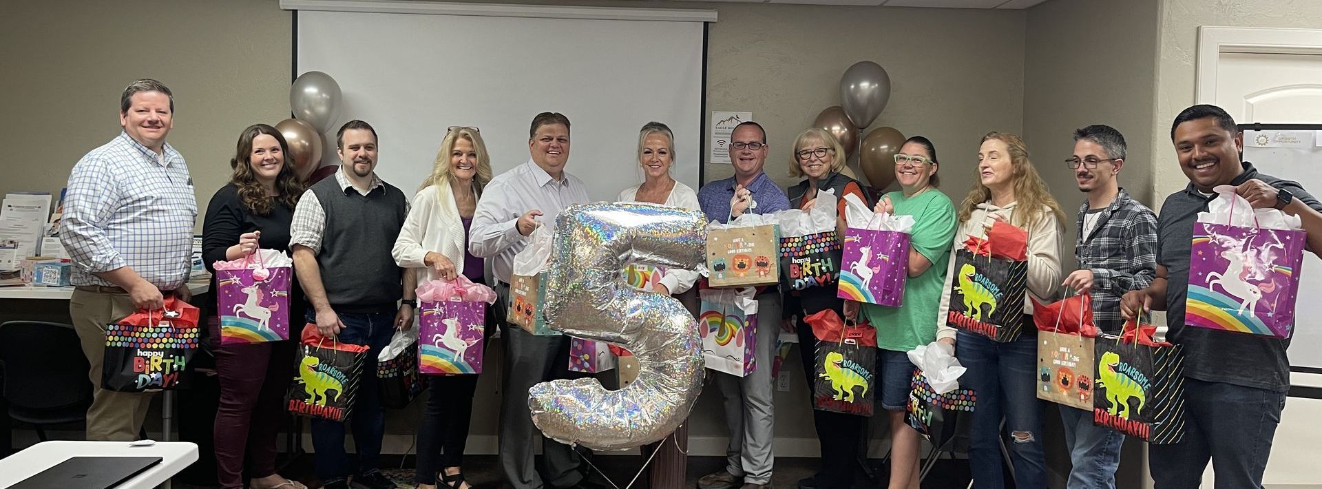 A group of people are standing in a room holding presents and balloons.