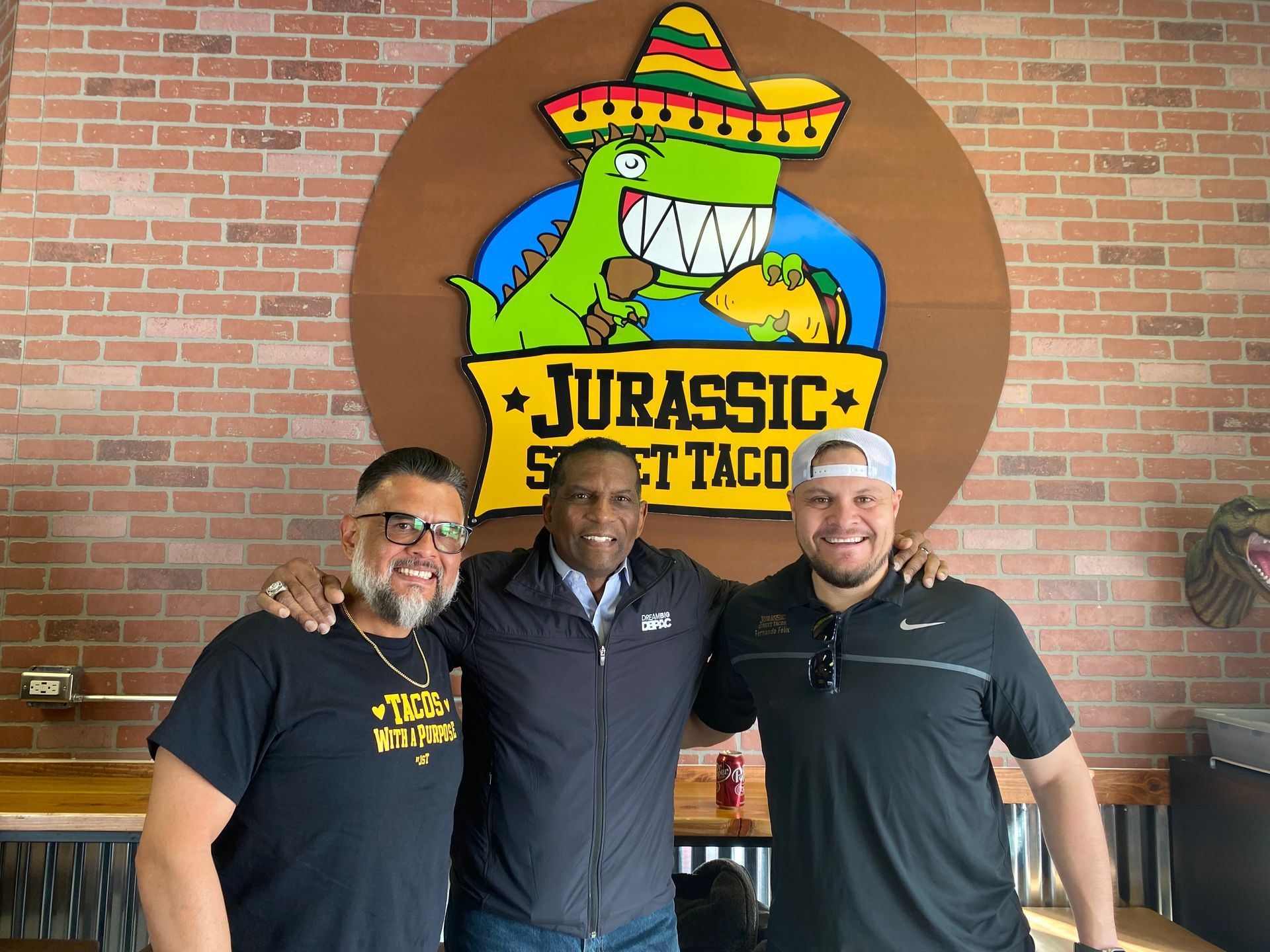 Three men are posing for a picture in front of a jurassic spot taco sign.