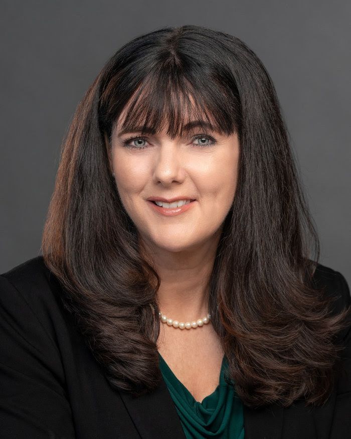 A woman with long hair and a pearl necklace is wearing a black jacket and a green shirt.