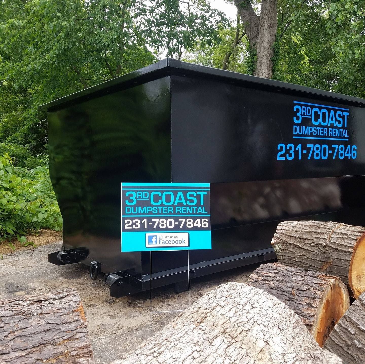 Black 3rd Coast dumpster on dirt, with firewood in front. Sign displays contact info.