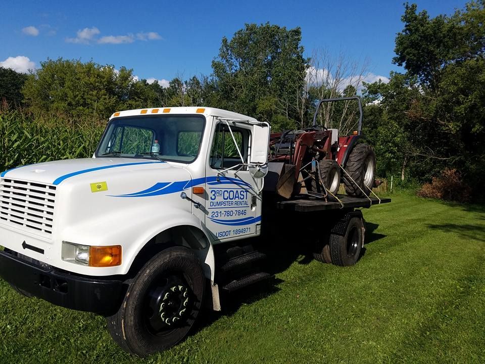 White truck with blue graphics hauling a small red tractor on a flatbed in a grassy field.