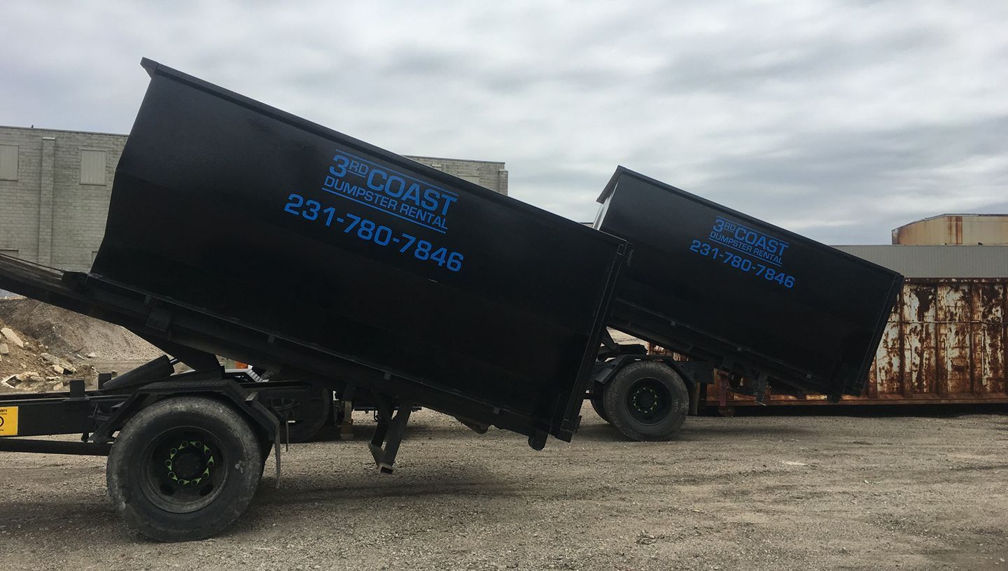 Two black roll-off dumpsters on trailers, tilted upwards. Gray gravel ground and cloudy sky.