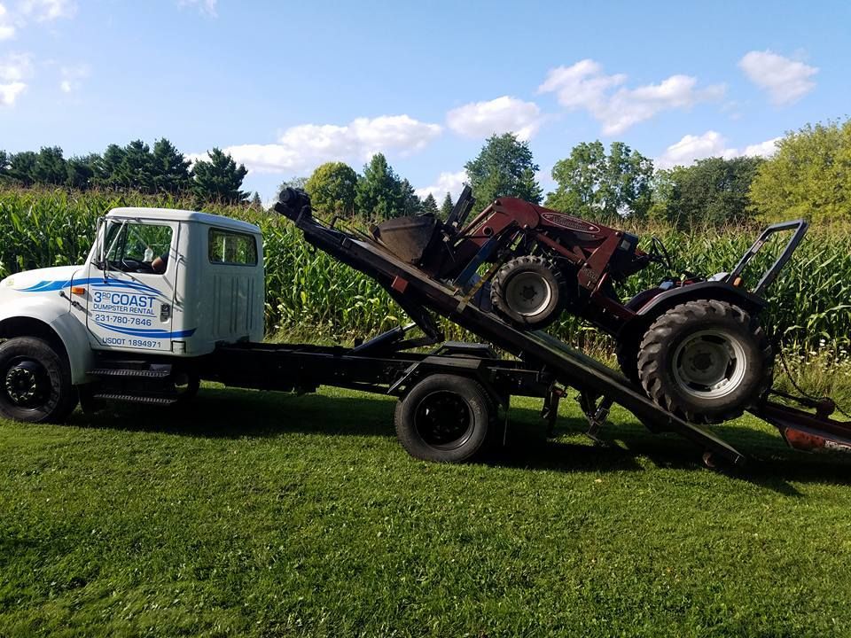 Tow truck with a red tractor on its bed, parked on a grassy area next to a cornfield.