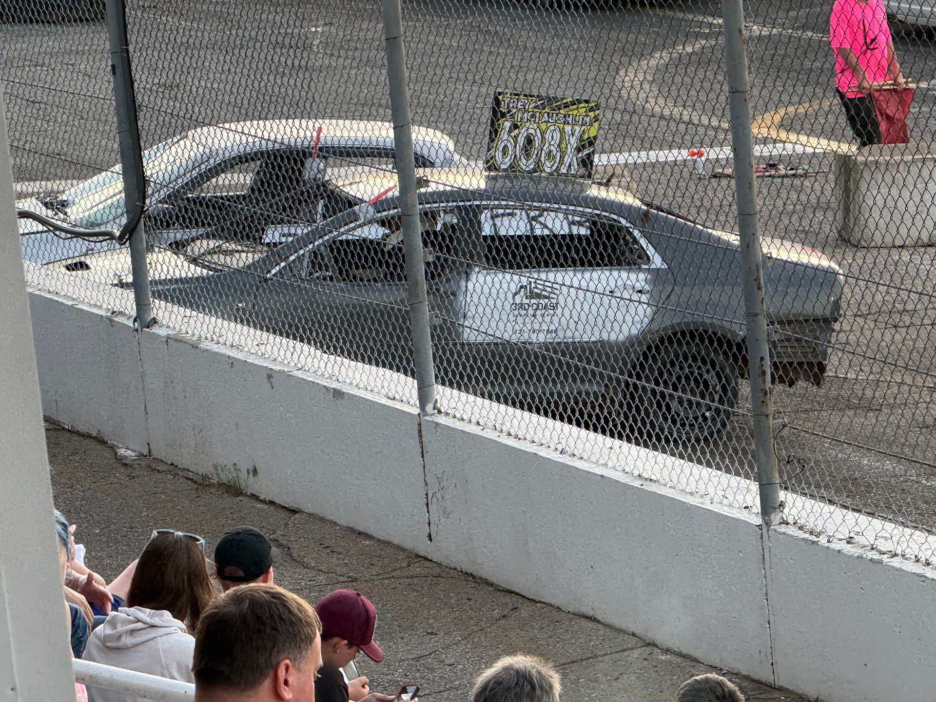 Gray race car on a dirt track behind a fence, with spectators watching from the stands.