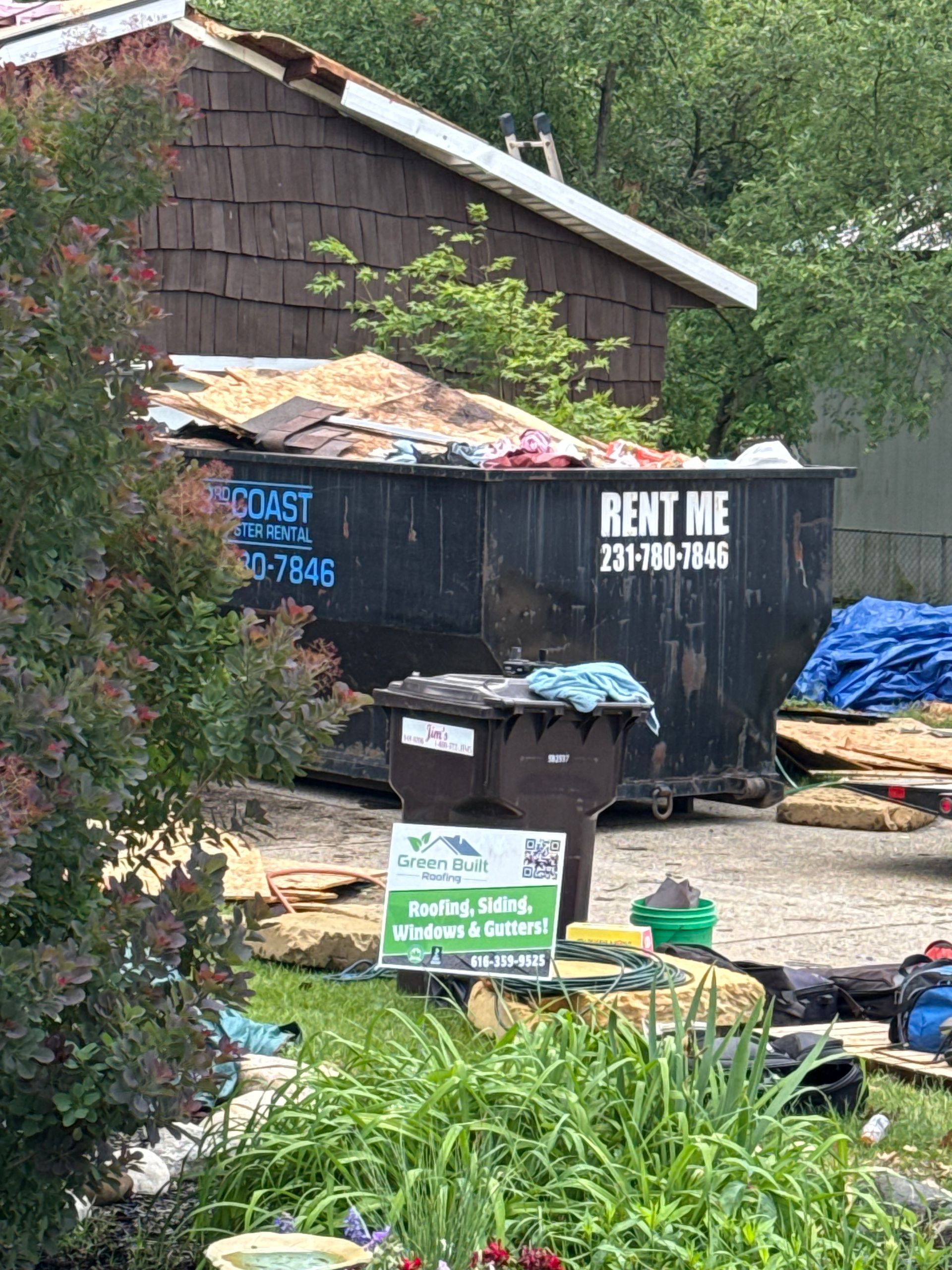 Dumpster beside house with a “RENT ME” sign, trash bags, and yard debris in a driveway