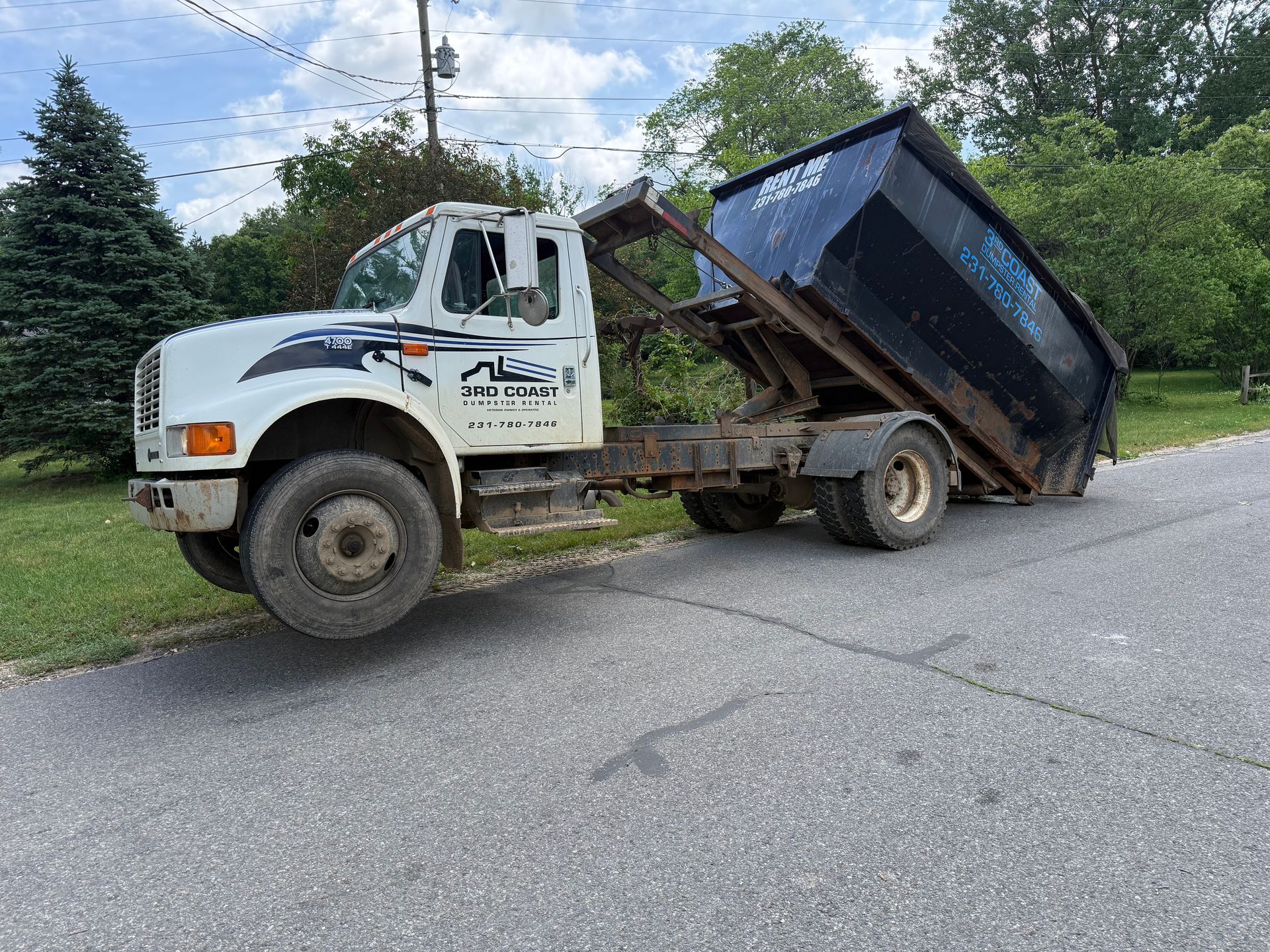 White dump truck with raised bed on a roadside, unloading or tipping debris into the grass.