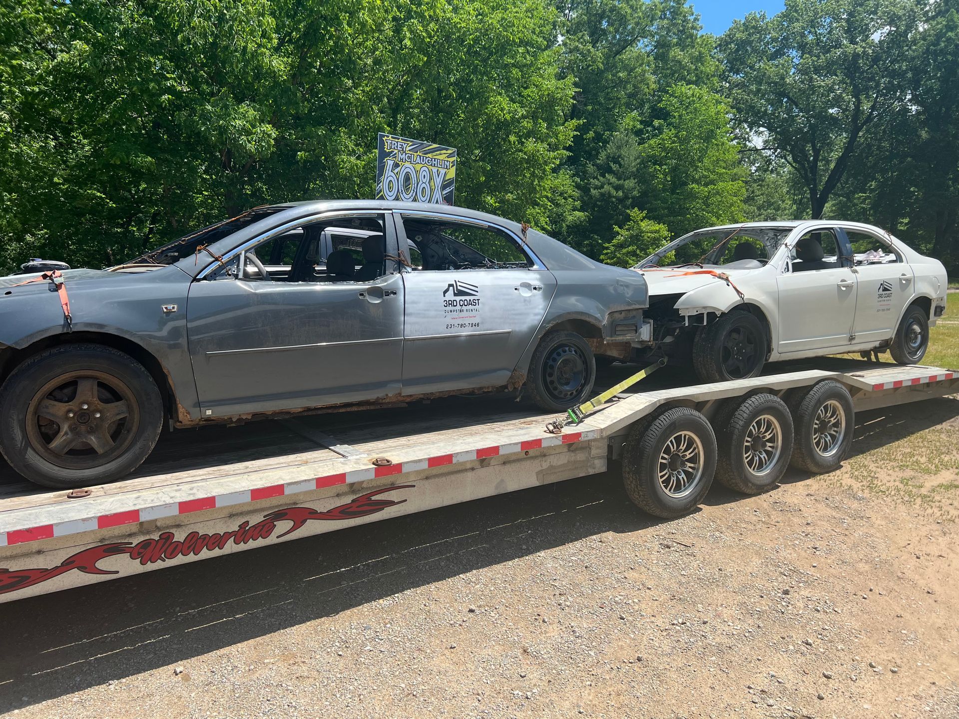 Two damaged cars on a trailer, a gray coupe behind a white sedan, beside a wooded roadside