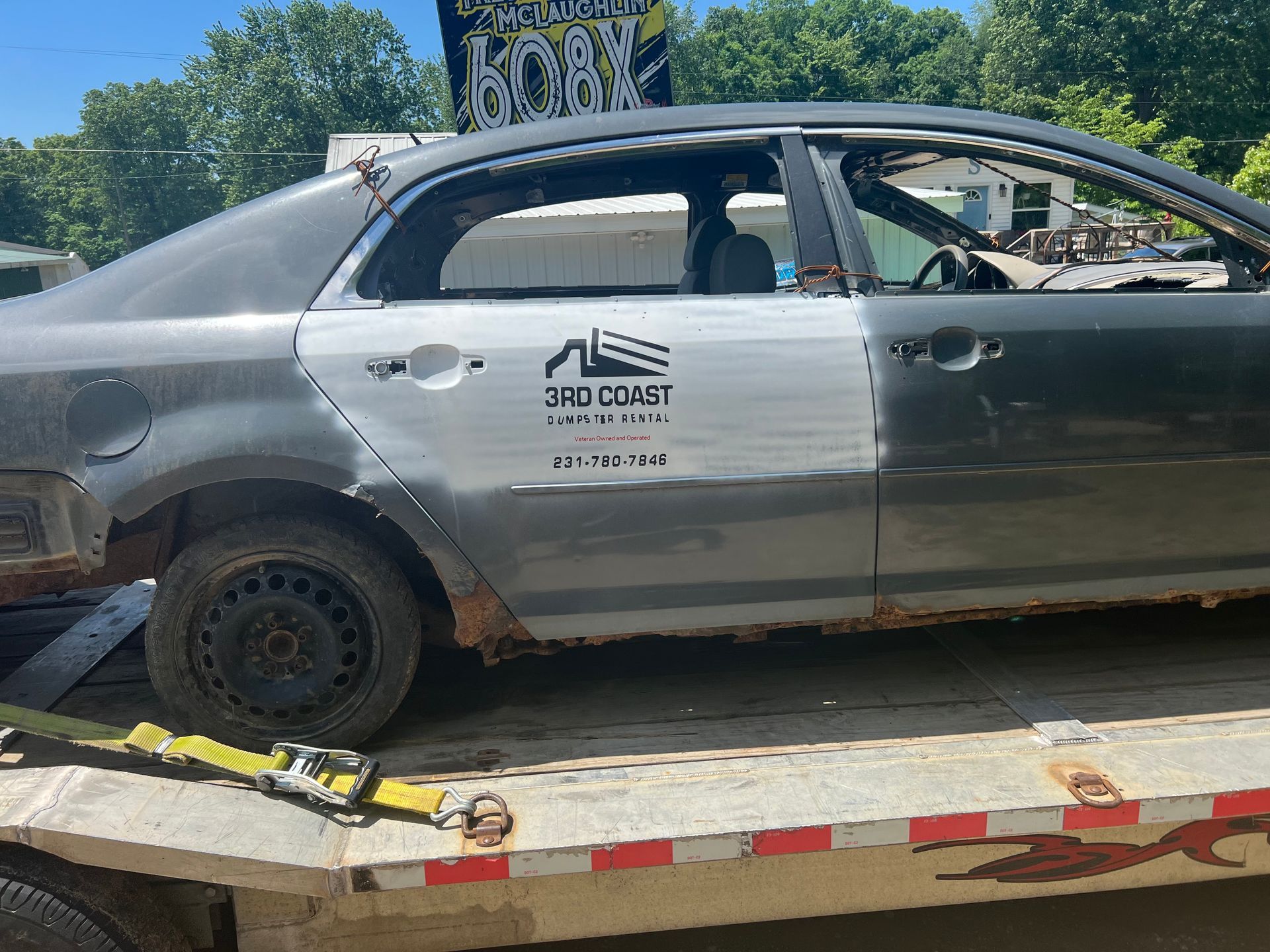 Damaged gray sedan on a tow trailer, missing windows and wheels, outdoors in daylight.